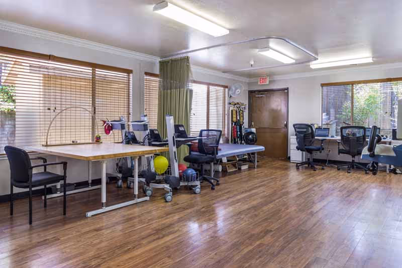 A spacious room with wooden flooring and large windows covered with blinds. The room contains several office chairs, tables with computer monitors, and physical therapy equipment including exercise balls and a treatment table. There is a green privacy curtain and an exit door at the back.
