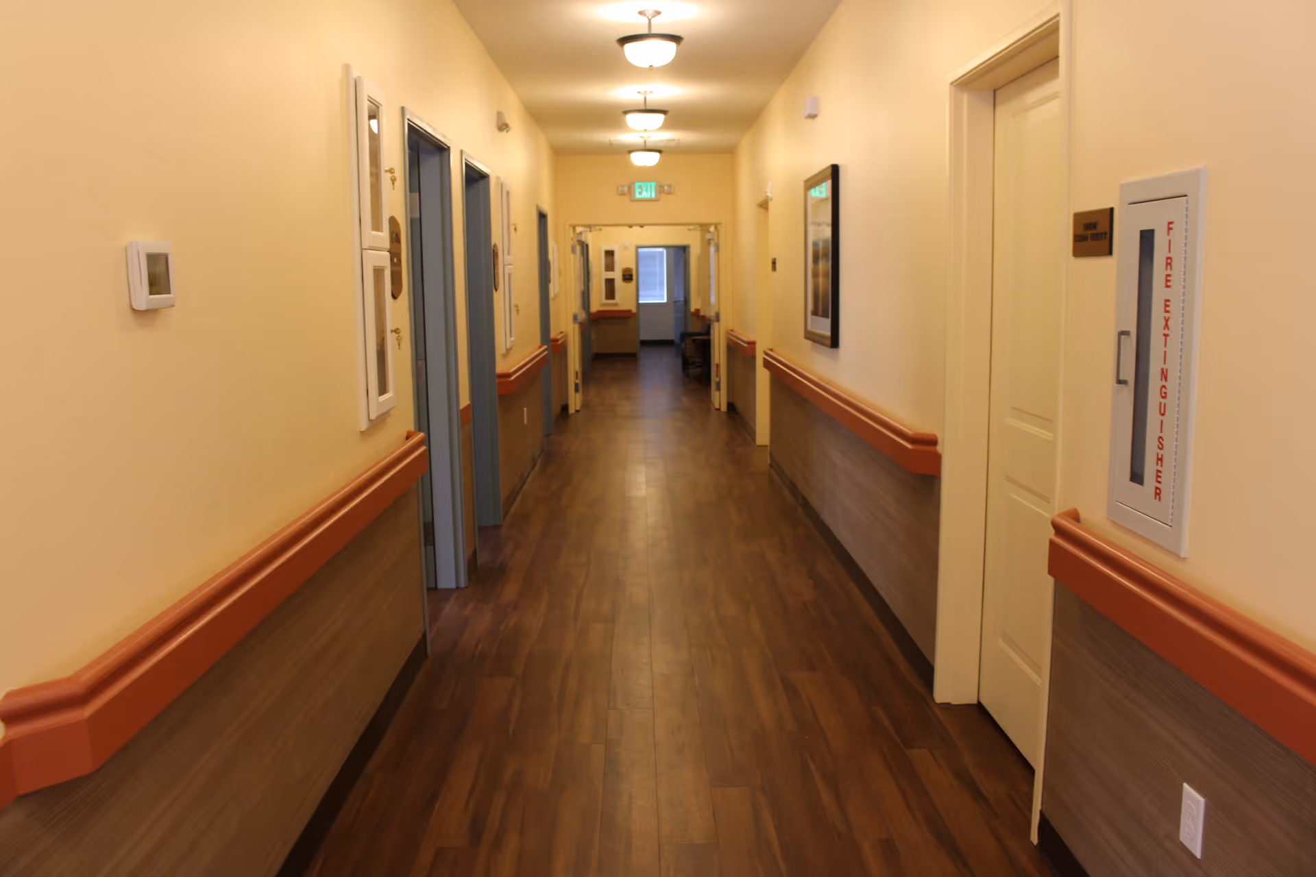 A long, well-lit hallway in a senior living facility with wooden flooring and beige walls. The hallway has multiple doors on both sides, handrails along the walls, ceiling lights, a fire extinguisher cabinet, and an exit sign at the far end.