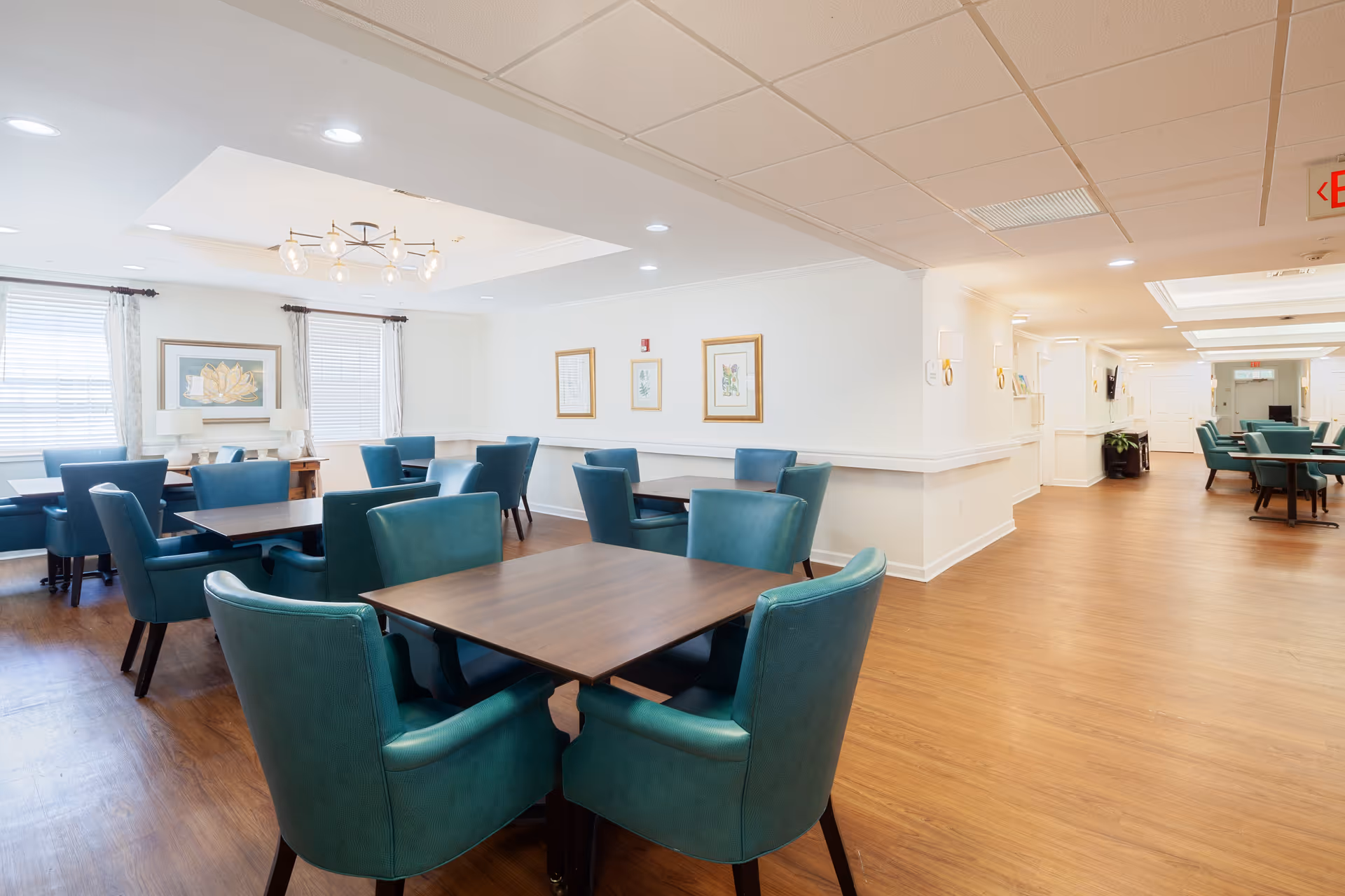 A bright and spacious dining room with multiple wooden tables surrounded by teal upholstered chairs. The room has large windows with white curtains, framed artwork on the walls, and a modern light fixture on the ceiling. The floor is wood, and the space extends into a hallway with more seating visible in the background.