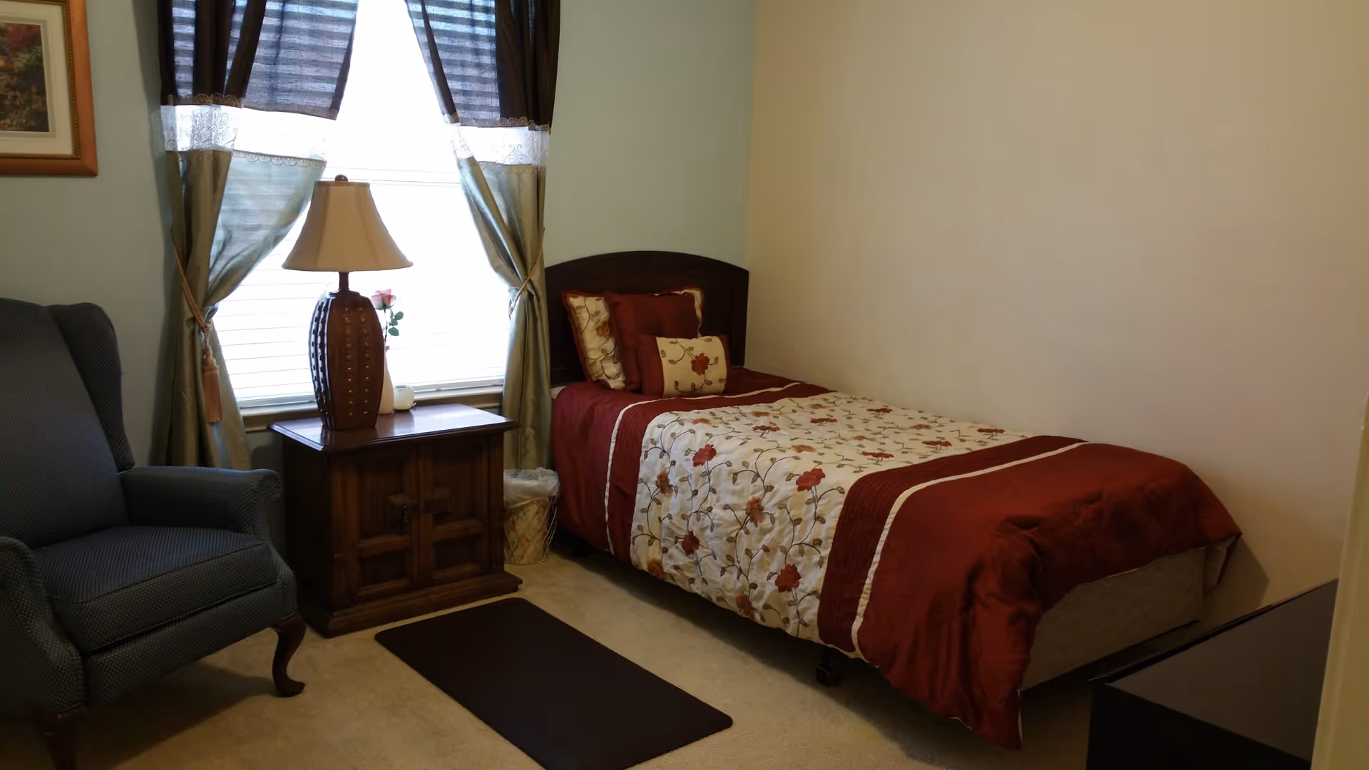Small bedroom with a single bed dressed in a red and floral comforter, a wooden nightstand and lamp by a curtained window, and an upholstered armchair.