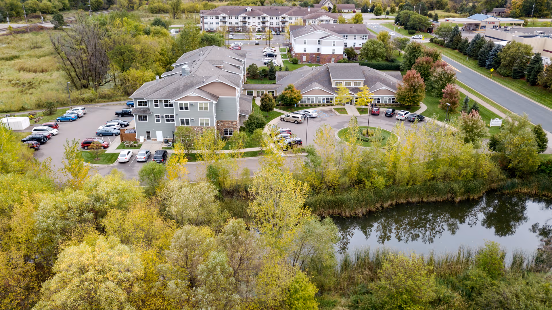 Aerial view of Legacy of Delano Senior Living facility surrounded by trees and greenery. The image shows multiple buildings with parking lots, a circular driveway, and a small pond in the foreground. The facility is located near a road with additional buildings visible in the background.