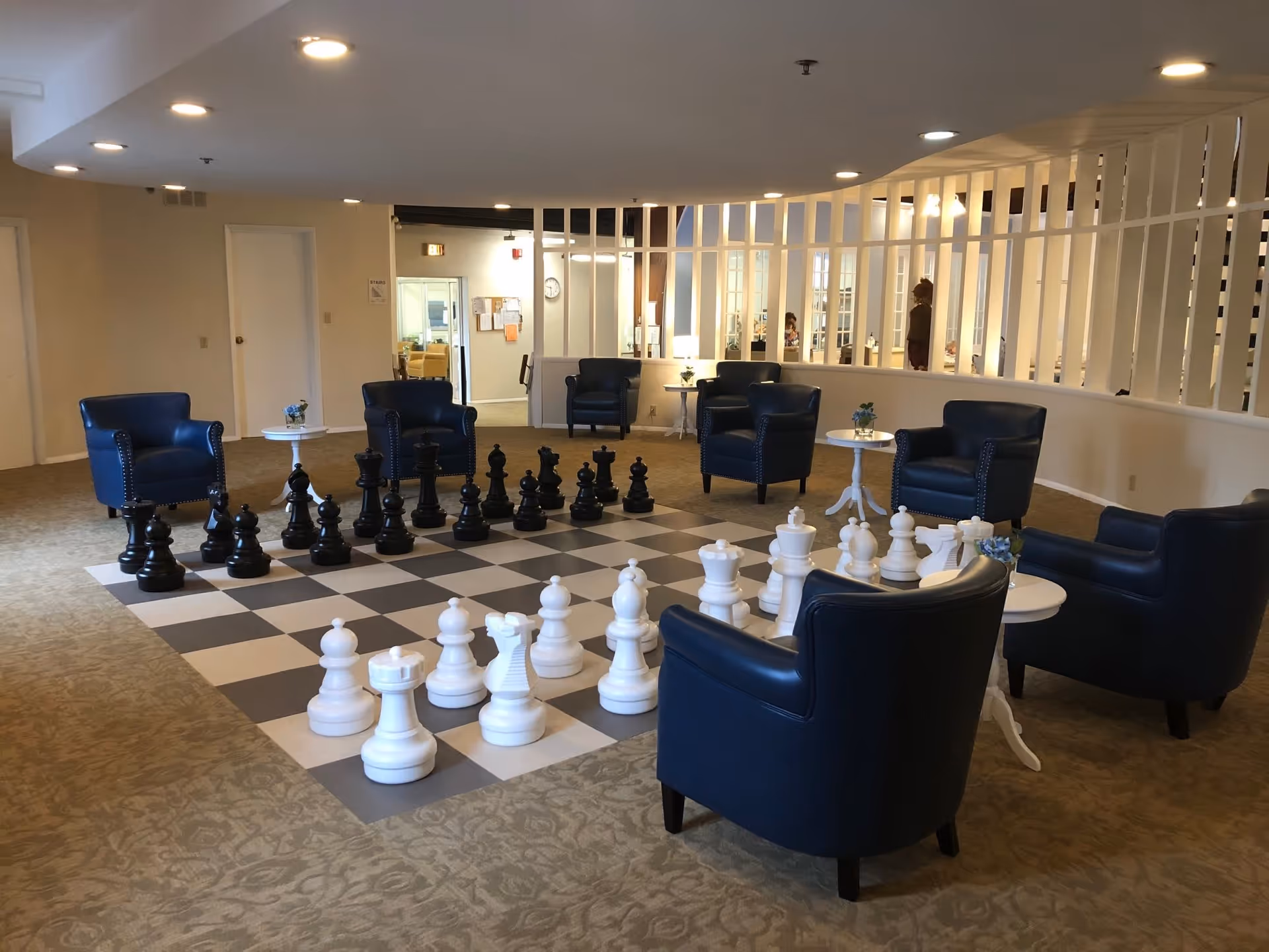 A spacious common area with a large floor chessboard and oversized chess pieces set up for a game. Surrounding the chessboard are several blue leather armchairs and small white side tables with flower arrangements. The room has beige carpeting, white walls, and recessed ceiling lights. A white partition with vertical slats separates this area from an adjacent room.