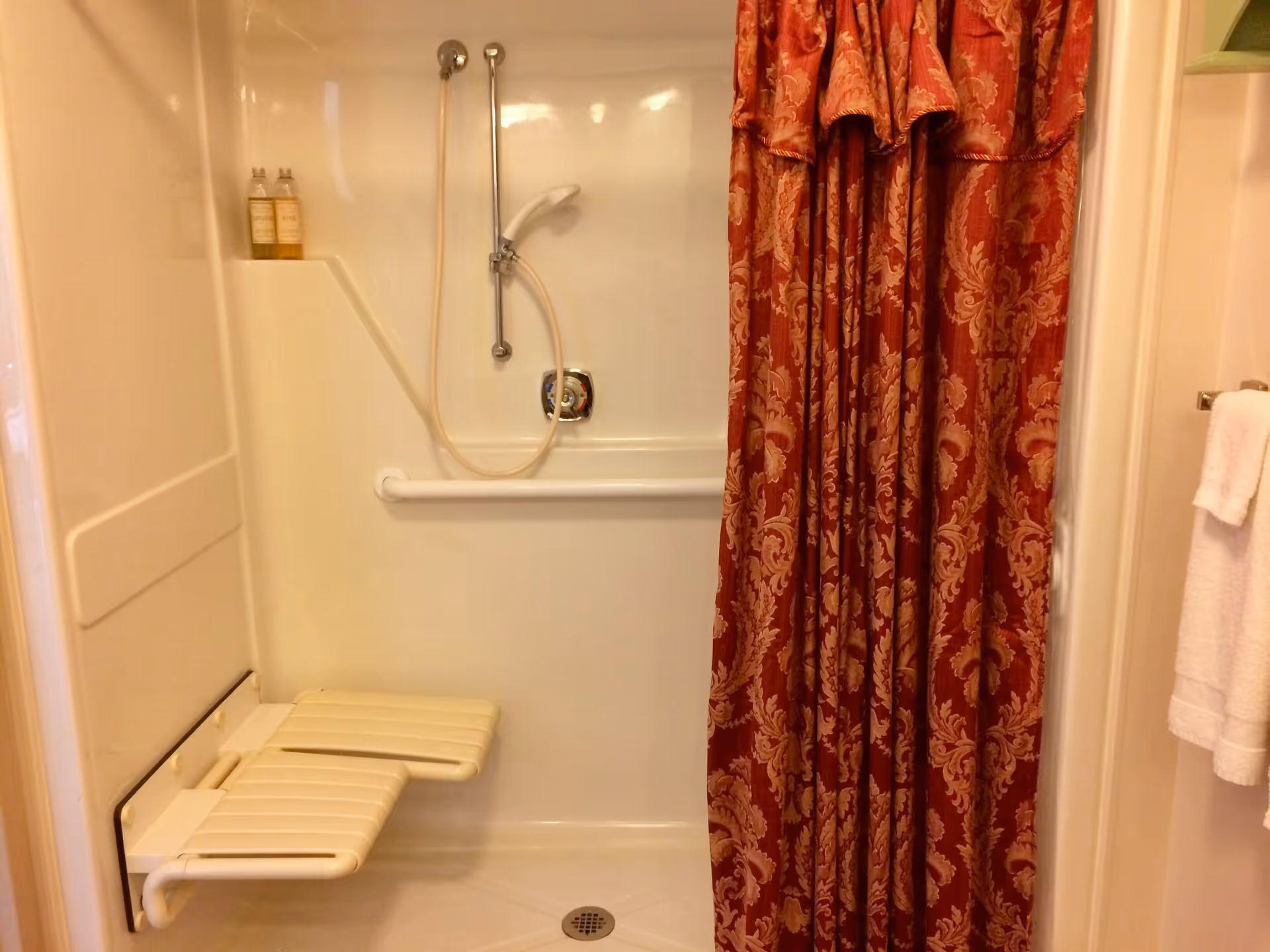 A bathroom shower area with a fold-down seat attached to the wall, a handheld showerhead on a sliding bar, two bottles of shower products on a built-in shelf, a grab bar for support, and a red patterned shower curtain partially drawn to the side. A white towel hangs on a towel rack outside the shower.