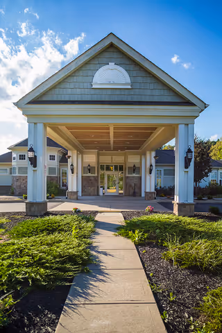Front entrance of a senior living facility with a covered driveway supported by white columns, surrounded by landscaped greenery and a clear blue sky with some clouds.