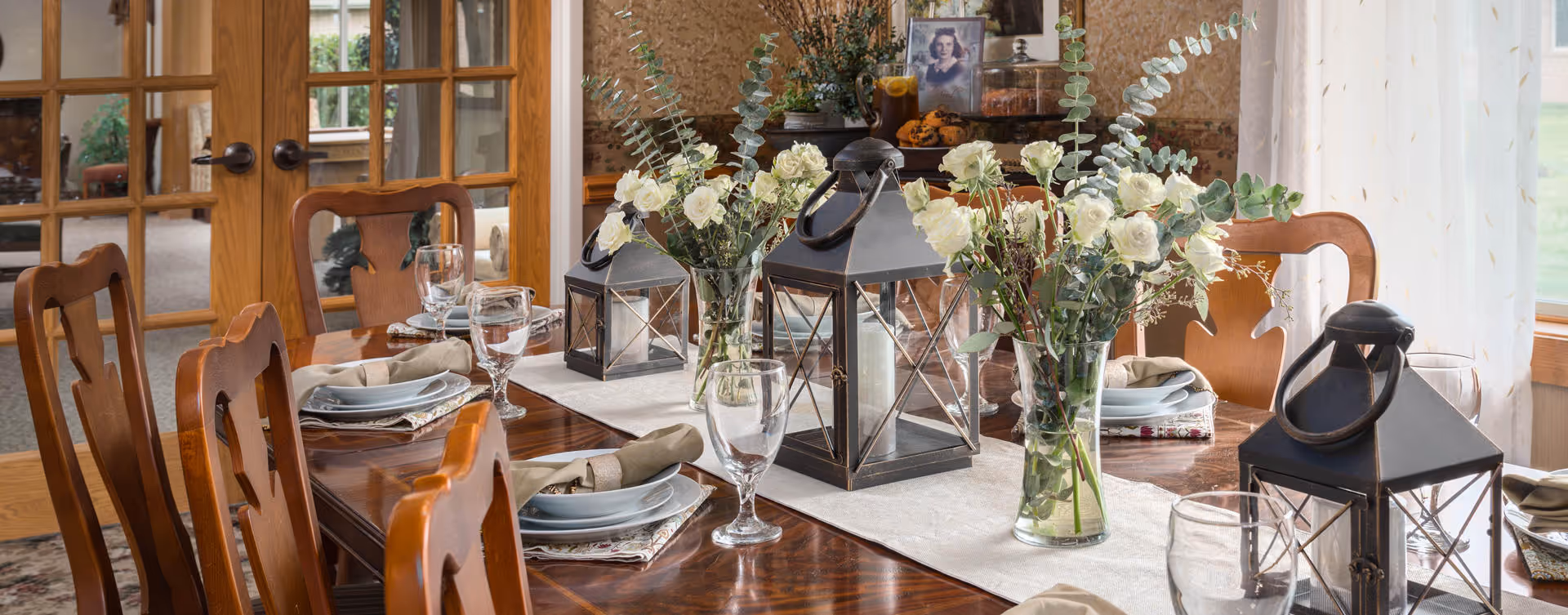 A dining table set with plates, napkins, and glasses, decorated with vases of white flowers and black lanterns. The room has wooden chairs and glass-paneled double doors in the background, with light curtains on the windows.