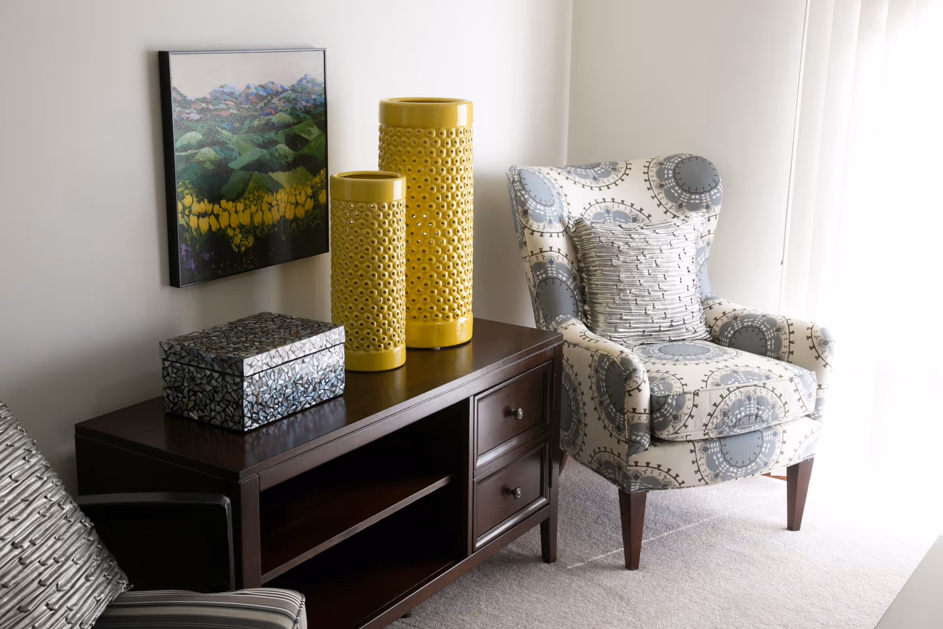 A cozy corner of a living room featuring a patterned armchair with a matching pillow, a dark wooden side table with two yellow decorative vases and a mosaic box, and a framed landscape painting on the wall. Light filters in through a window with sheer white curtains.