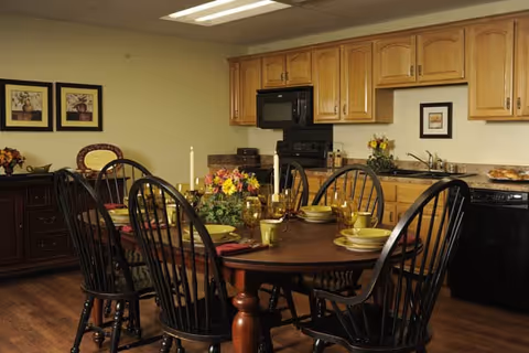 Dining area with a wooden table set for a meal and black spindle-back chairs in front of a kitchen with wood cabinets and appliances.