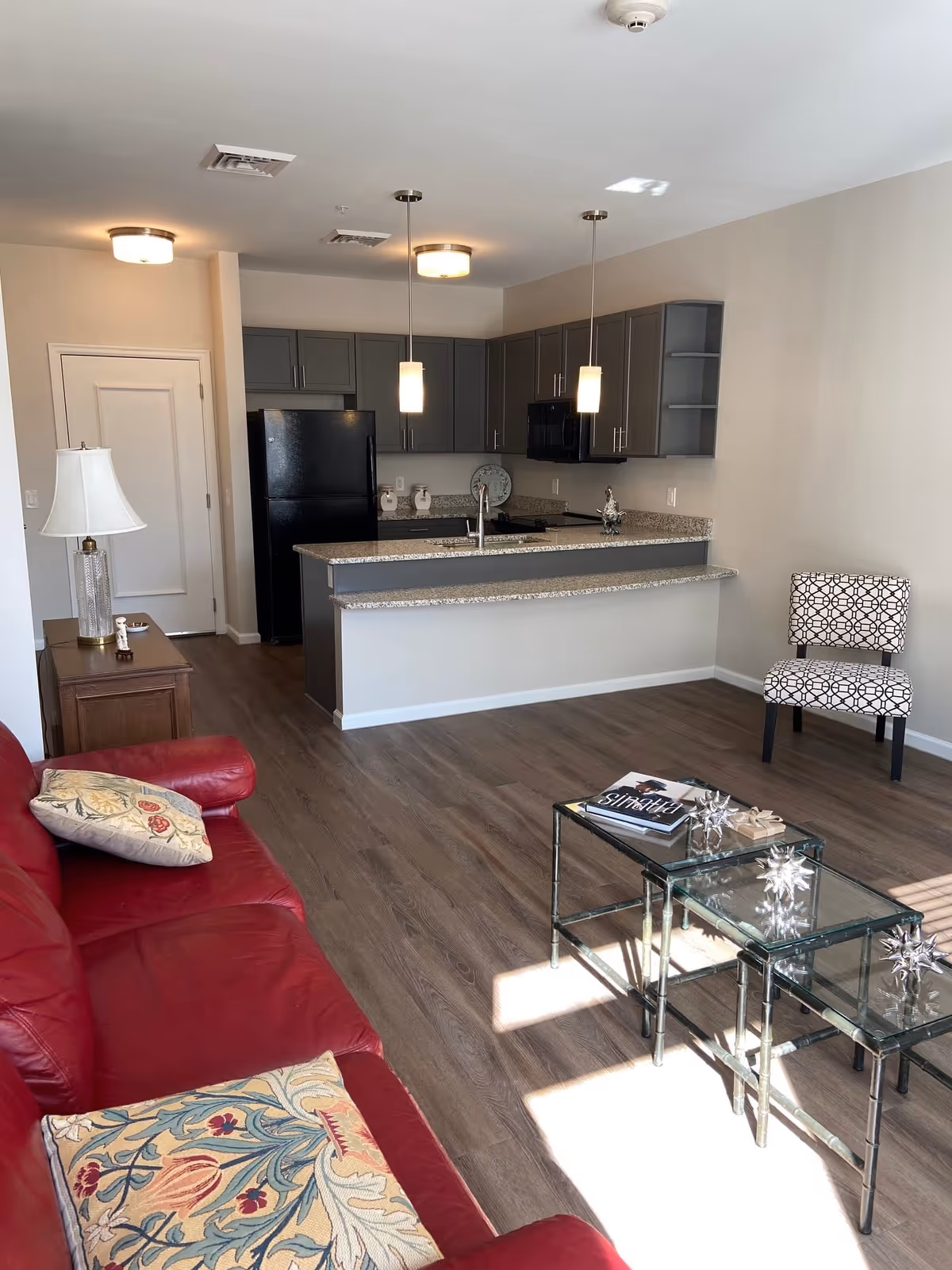A modern living area with a red leather sofa adorned with patterned cushions, a glass coffee table with decorative items and a magazine, a patterned chair against the wall, and an open kitchen with gray cabinets, a black refrigerator, a granite countertop, and two pendant lights hanging above the kitchen island.