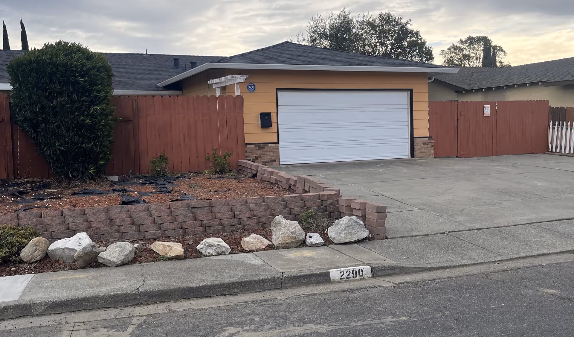 A residential driveway with a closed white garage door attached to a single-story house. The house has a yellow exterior with brick accents and a red wooden fence surrounding the property. There is a small landscaped area with rocks and a bush next to the driveway. The house number 2290 is painted on the curb.