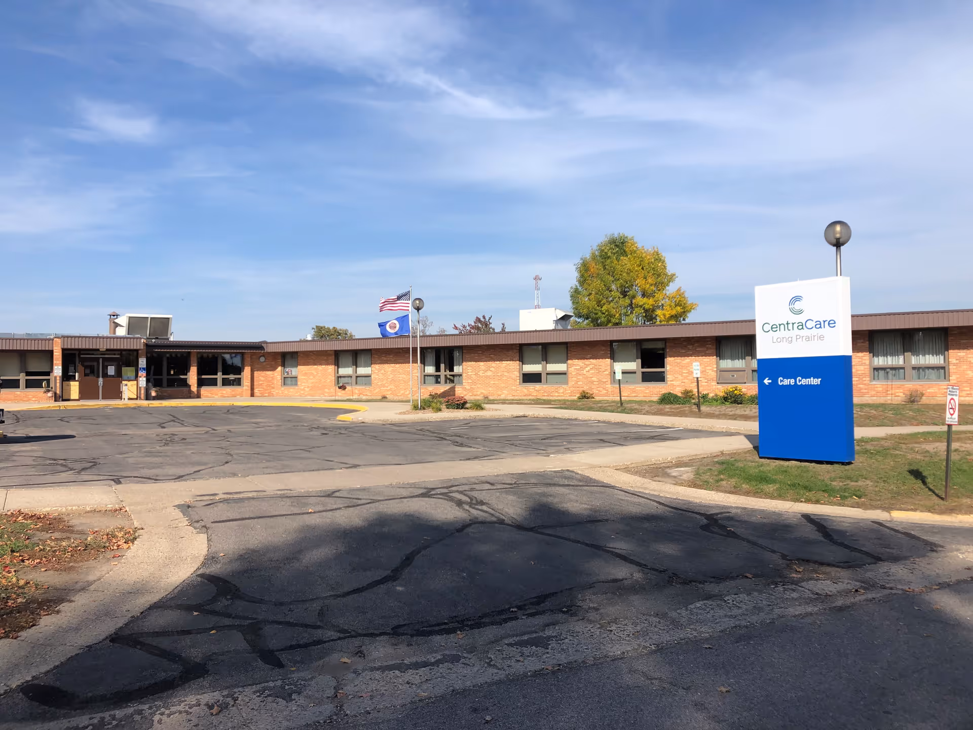 Exterior view of the Long Prairie Care Center building with a parking lot in front, two flagpoles with the American and Minnesota state flags, and a sign that reads 'CentraCare Long Prairie Care Center' on the right side of the image under a clear blue sky.