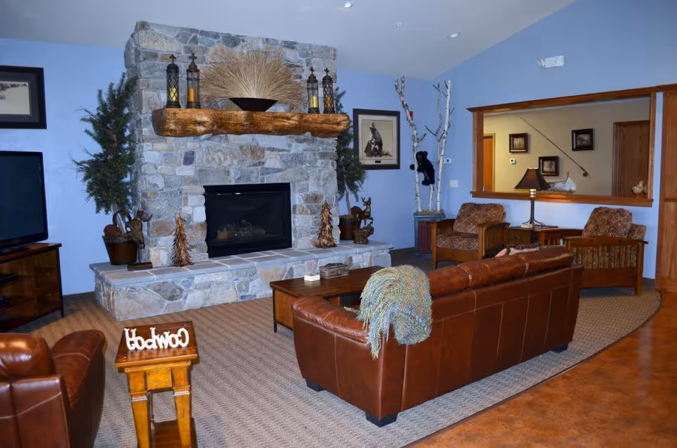 Cozy living room with a stone fireplace featuring a wooden mantle decorated with lanterns and a bowl of dried plants. The room has light blue walls, a brown leather sofa with a knitted throw, two patterned armchairs, a wooden coffee table, and a TV on a wooden stand. There are decorative plants and framed artwork on the walls.