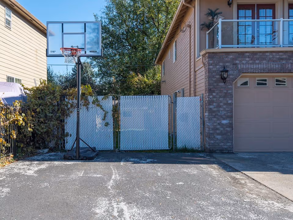 Outdoor area showing a portable basketball hoop on a paved surface next to a brick building with a garage door. There is a white chain-link fence with some greenery behind it and trees in the background under a clear blue sky.