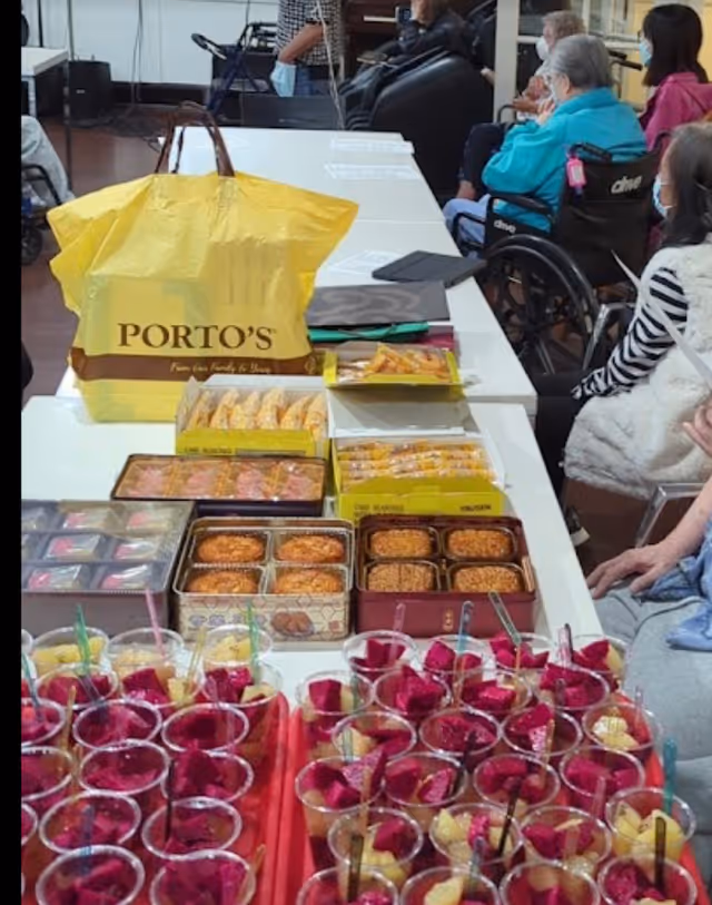 A table filled with various packaged snacks and cups of fruit, including dragon fruit and pineapple, in a room where several elderly people are seated, some in wheelchairs, wearing masks.