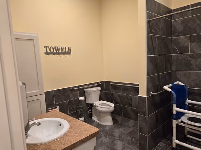 Accessible bathroom with a sink and countertop, toilet, tiled walk-in shower with a blue shower chair and grab bars, and a wall sign that says 'TOWELS'.