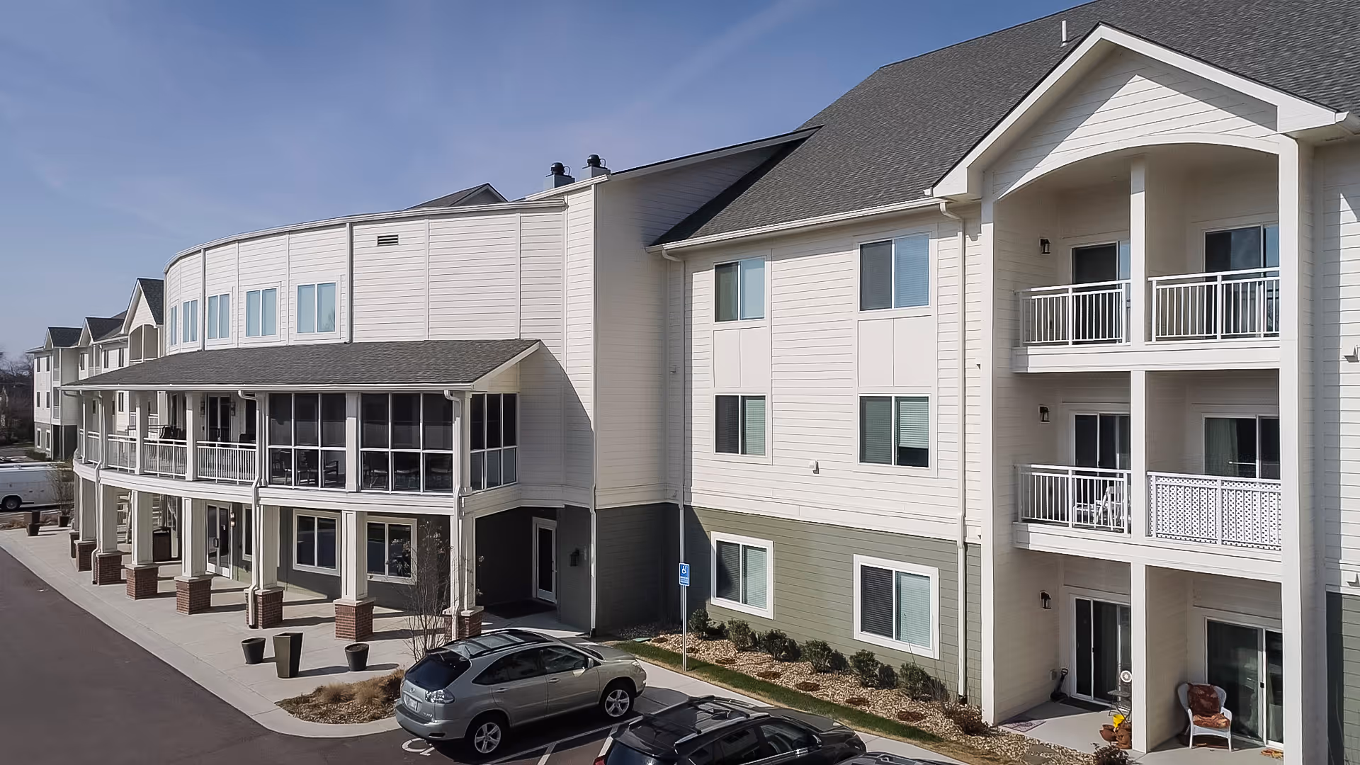 Exterior view of a multi-story senior living facility building with balconies and a covered porch area. There are a few parked cars in front and landscaping along the building's base under a clear sky.