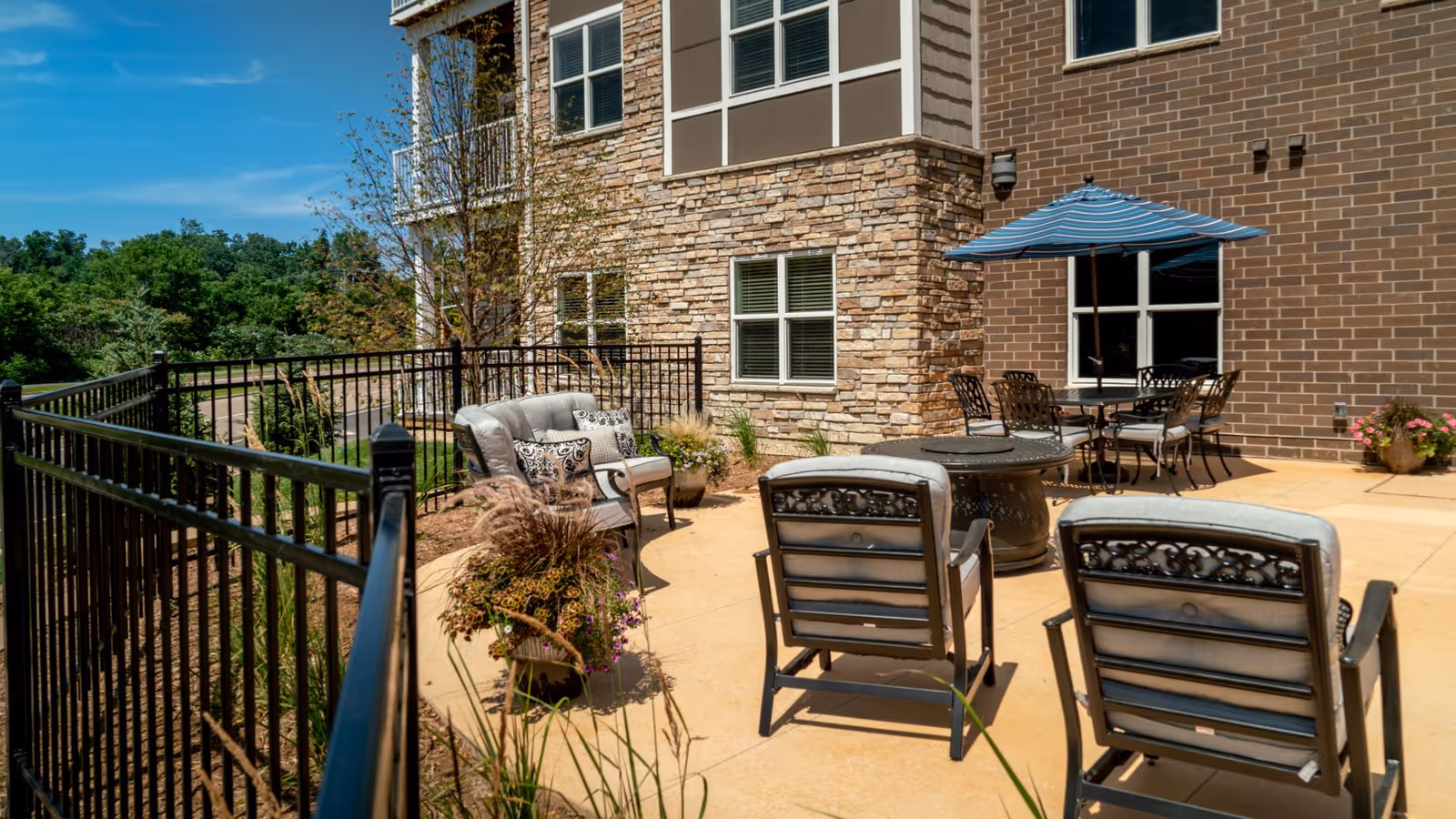 Outdoor patio area at Prairie Bluffs Senior Living with cushioned chairs, a round table with a blue umbrella, potted plants, and a black metal fence surrounding the space. The building exterior features stone and brick walls with windows.