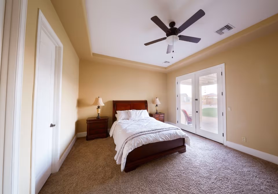 A bedroom with beige walls and carpeted floor featuring a wooden bed with white bedding and pillows. There are two matching wooden nightstands with lamps on either side of the bed. A ceiling fan with lights is mounted on the ceiling. French doors lead outside to a patio area with outdoor chairs visible through the glass.