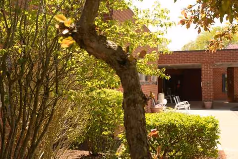 Front exterior of a brick senior care facility with greenery and white seating near entrance.