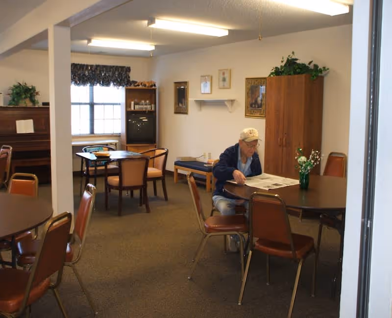 A senior man wearing a cap and jacket is sitting at a round table reading a newspaper in a common room. The room has several round tables with chairs, a piano, a TV on a stand, framed pictures on the walls, and a wooden cabinet with a plant on top. There is a window with a valance letting in natural light.