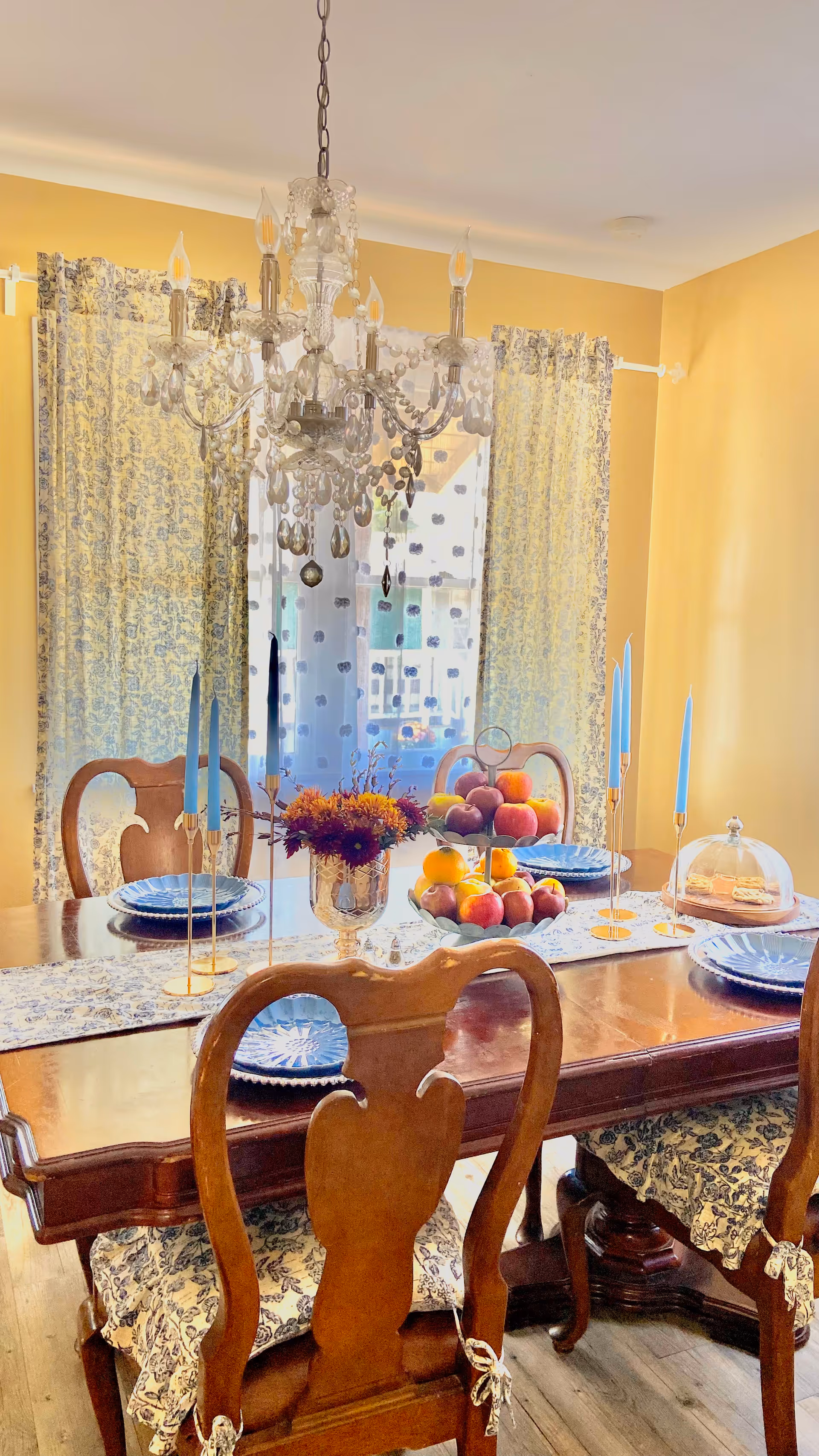 A warmly lit dining room with a wooden table set for four. The table features blue plates, tall blue candles in gold holders, a centerpiece with a bouquet of flowers, and a tiered fruit stand filled with apples and oranges. The chairs have wooden backs and patterned cushions. A crystal chandelier hangs above the table, and floral curtains cover the window in the background.