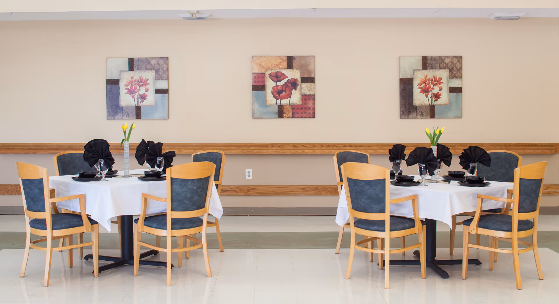 Two round dining tables set with white tablecloths, black napkins folded in a decorative manner, black plates, and wine glasses. Each table has a vase with yellow tulips. The tables are surrounded by wooden chairs with blue cushions. The background features a beige wall with three framed floral paintings.