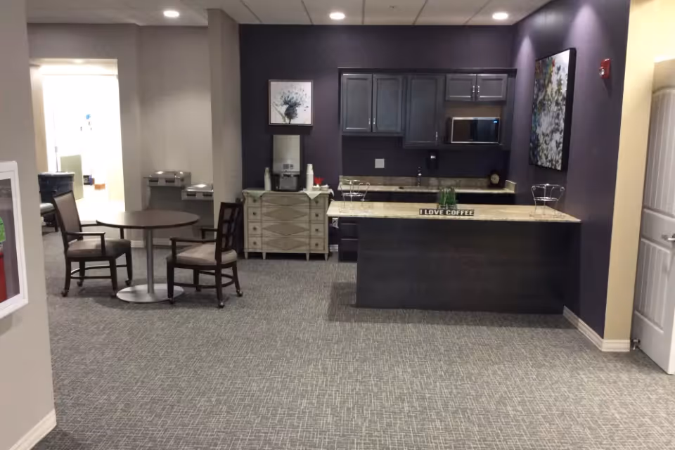 Interior view of a common area in a senior living facility named Serenity, featuring a small kitchenette with dark cabinets, a countertop with a sign that reads 'I LOVE COFFEE', a coffee machine on a side cabinet, two chairs around a round table, and artwork on the walls.