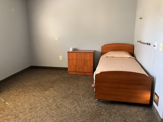 A simple room with a single wooden bed with beige bedding and a matching wooden dresser against a light gray wall. The floor is carpeted in a brown pattern, and the room appears minimally furnished.