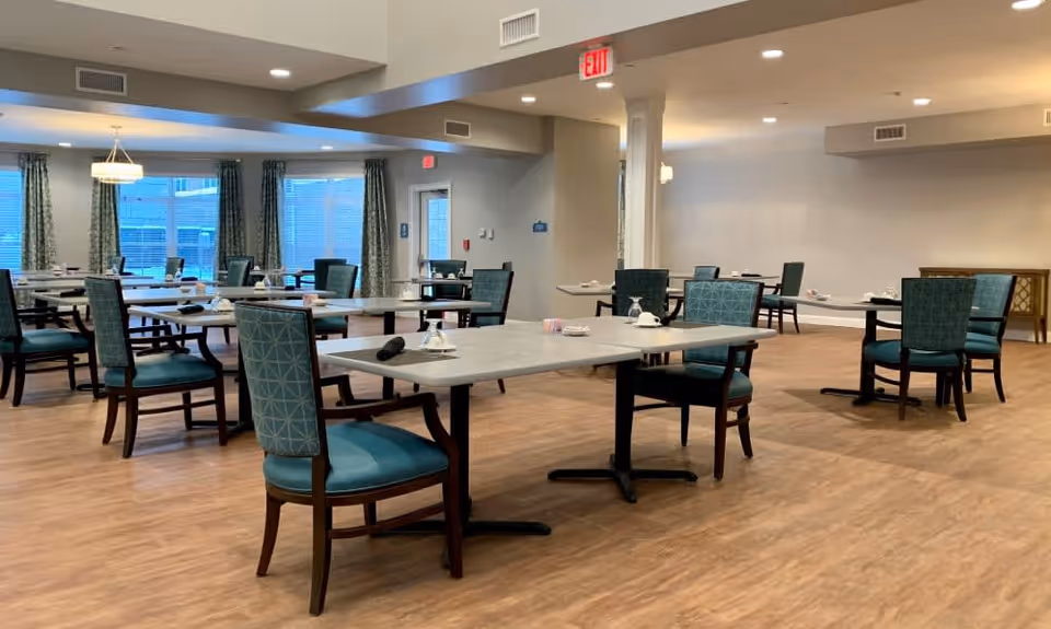 A spacious dining room with multiple tables and chairs arranged neatly. Each table has place settings including cups, saucers, and napkins. The room has large windows with curtains, light wood flooring, and neutral-colored walls with ceiling lights providing bright illumination.
