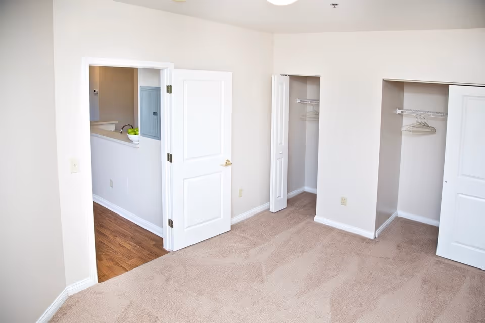Empty room with beige carpet, two open closets with hangers, and a white door leading to a hallway with wooden flooring and a bowl of green apples on a counter.