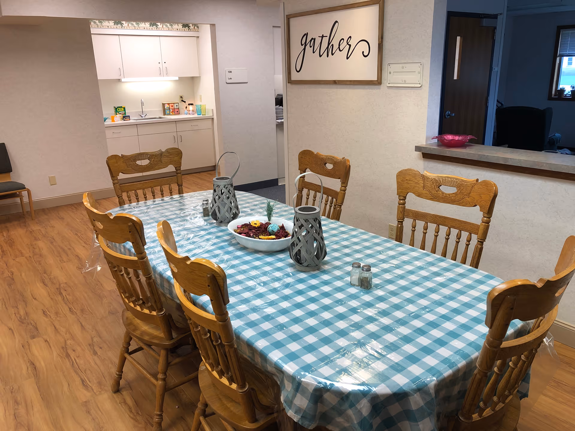 Long dining table with a blue-and-white checkered tablecloth surrounded by wooden chairs in a communal dining area with a kitchenette and a 'gather' sign on the wall.