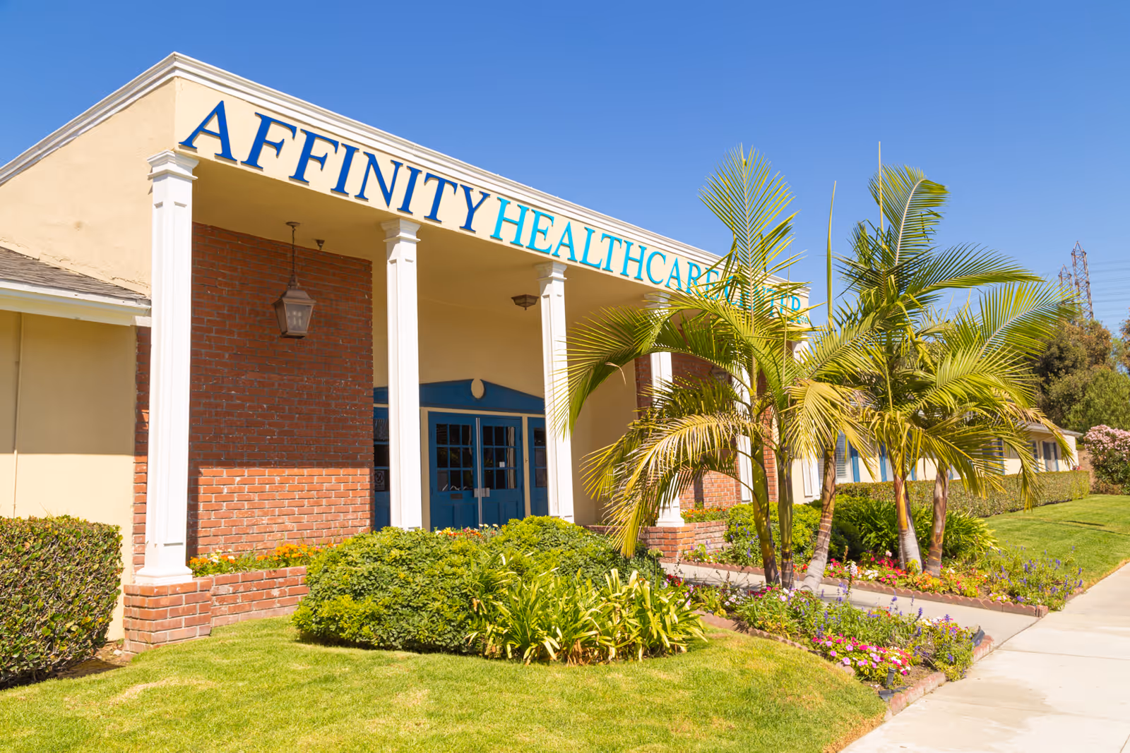 Exterior view of a healthcare facility building with a sign reading 'AFFINITY HEALTHCARE' above the entrance. The building has brick and beige walls, white columns, blue double doors, and is surrounded by green bushes, palm trees, and a well-maintained lawn under a clear blue sky.