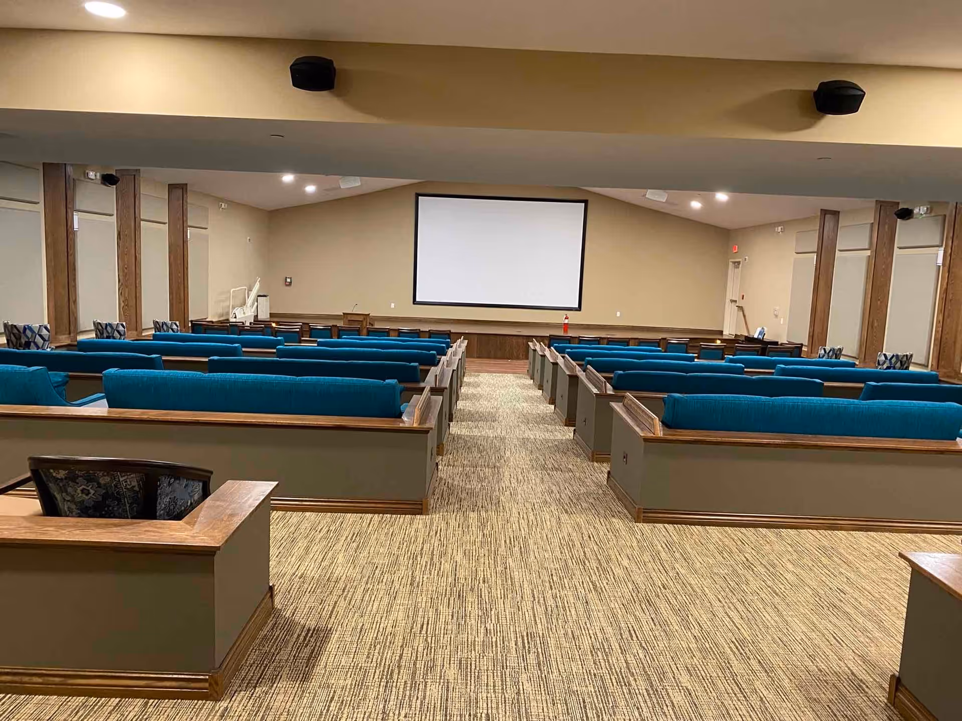 Interior view of a large meeting or presentation room with rows of blue cushioned benches facing a blank projection screen on the far wall. The room has beige walls, carpeted floor, wooden trim, and ceiling-mounted speakers.