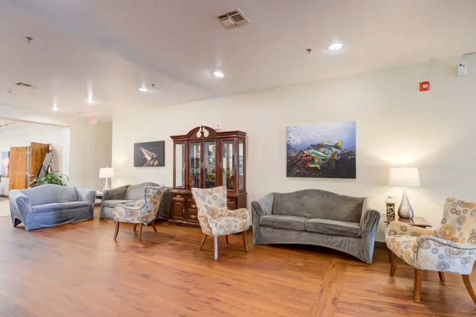 Spacious furnished lounge with gray sofas, patterned armchairs, a wooden display cabinet, artwork, and lamps on hardwood floors.