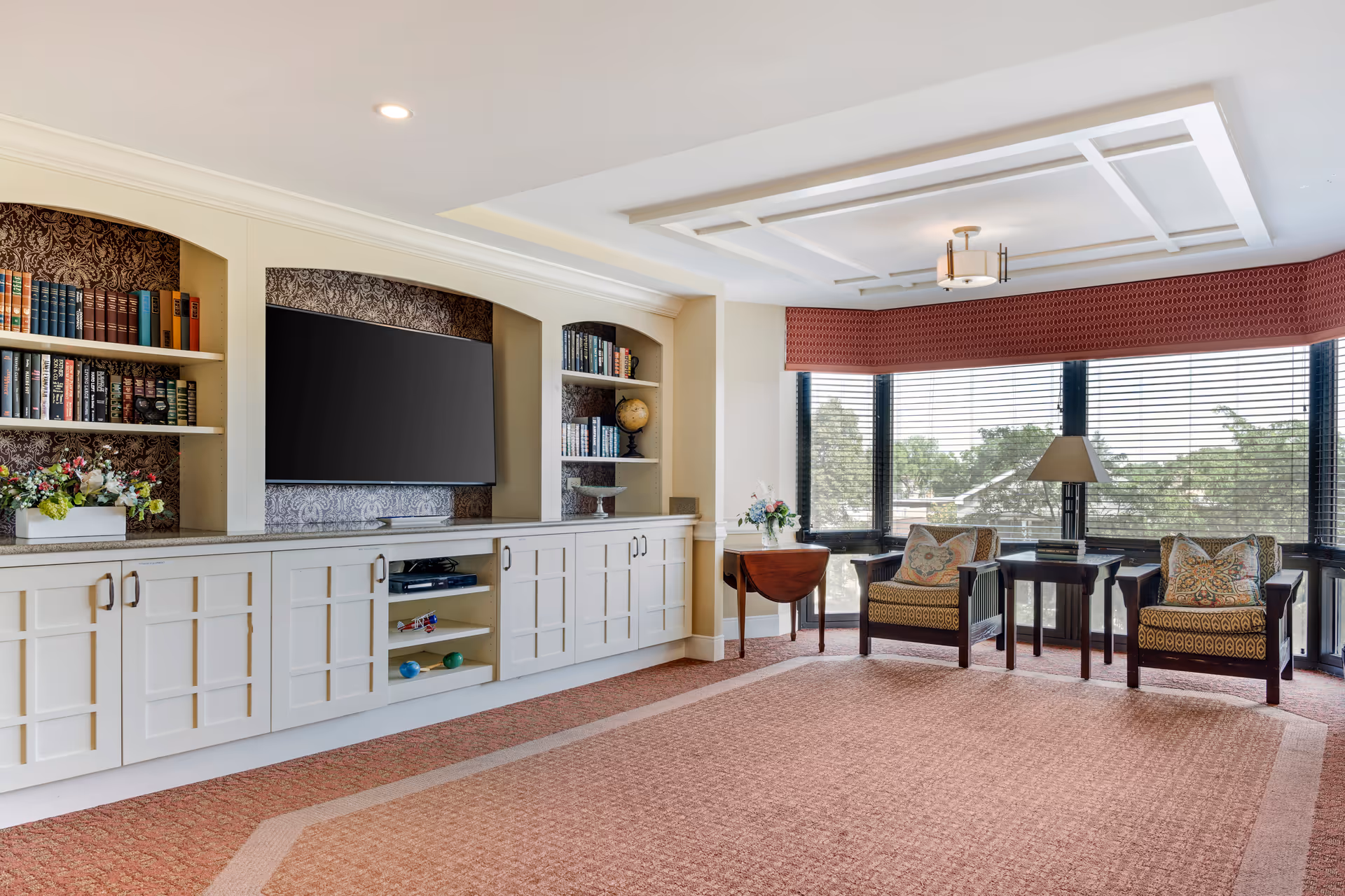 A bright and spacious living room with large windows covered by blinds, two patterned armchairs with decorative pillows, a side table with a lamp, and built-in white cabinetry with shelves holding books, a globe, and decorative items. A flat-screen TV is mounted in the center of the cabinetry. The room has a patterned carpet and a coffered ceiling with a modern light fixture.