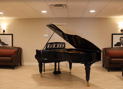 A black grand piano with its lid open is placed in the center of a room with beige tiled flooring and cream-colored walls. On either side of the piano, there are brown leather sofas with red patterned cushions. Above each sofa, there is a framed black and white portrait. The room is softly lit by wall sconces and ceiling lights.