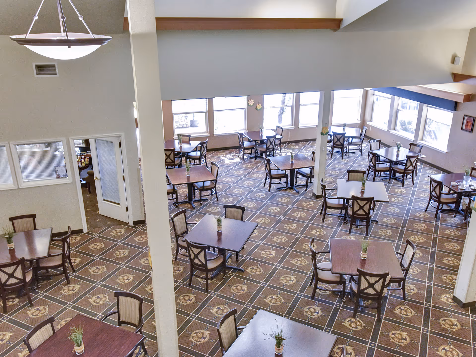 Spacious senior living dining room with multiple square tables and chairs arranged on patterned carpet and small potted plants on each table beneath large windows.