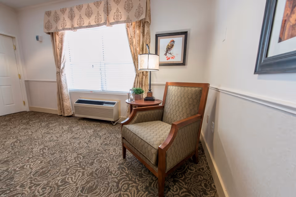 A cozy corner in a room with a patterned armchair next to a small round wooden table holding a lamp and a small plant. Behind the chair is a window with patterned curtains and a wall-mounted framed picture of a bird. The floor is covered with a patterned carpet.