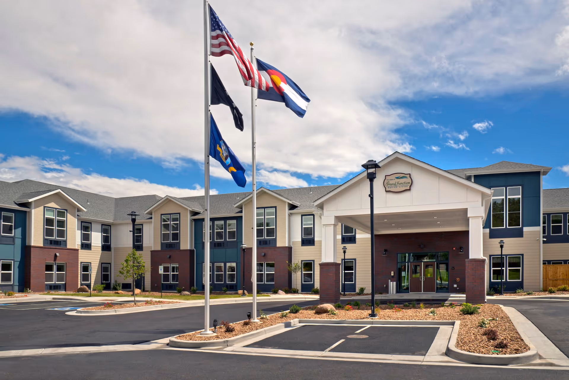 Exterior view of a two-story senior living facility with a covered entrance, multiple windows, and three flagpoles displaying the American flag, Colorado state flag, and another flag. The building has beige and blue siding with brick accents and a clear sky with some clouds in the background.
