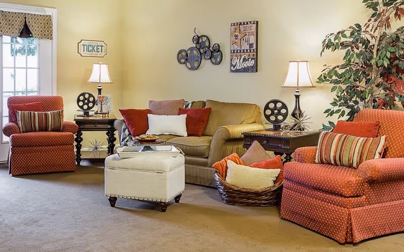 A cozy living room featuring two red patterned chairs, a beige sofa with colorful cushions, a coffee table, and decorative elements like lamps and wall art.