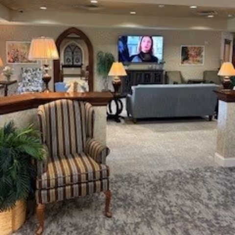 A cozy living room area in a senior living facility with a striped armchair in the foreground, a potted plant to the left, and a blue sofa facing a wall-mounted TV. The room is softly lit with table lamps and decorated with framed artwork and an arched doorway in the background.