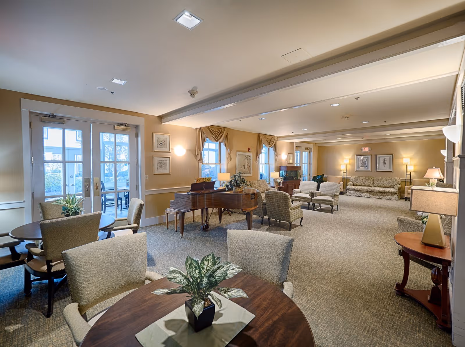 A spacious and well-lit common area in a senior living facility featuring multiple seating arrangements including armchairs and sofas. There is a grand piano in the center of the room, several tables with chairs, decorative plants, framed artwork on the walls, and large windows with drapes allowing natural light to enter. The room has a warm, inviting atmosphere with soft lighting from wall sconces and lamps.