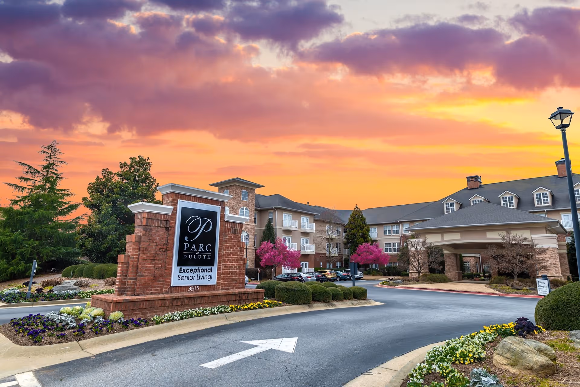 Entrance to Parc at Duluth senior living facility with a large brick sign displaying the facility name and slogan 'Exceptional Senior Living!' surrounded by landscaped flowers and bushes, with the building and parking area in the background under a colorful sunset sky.