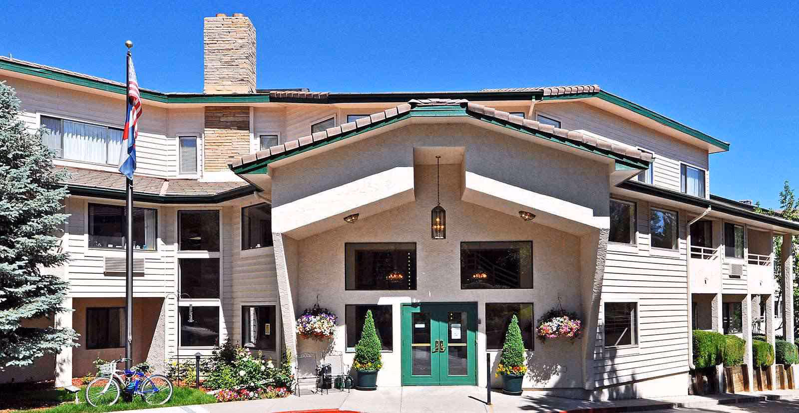 Exterior front view of a two-story senior living facility building with beige siding and a green roof trim. The entrance features green double doors under a covered porch with hanging flower baskets and potted plants. An American flag and another flag are displayed on a flagpole to the left, with a bicycle parked nearby. The sky is clear and blue.