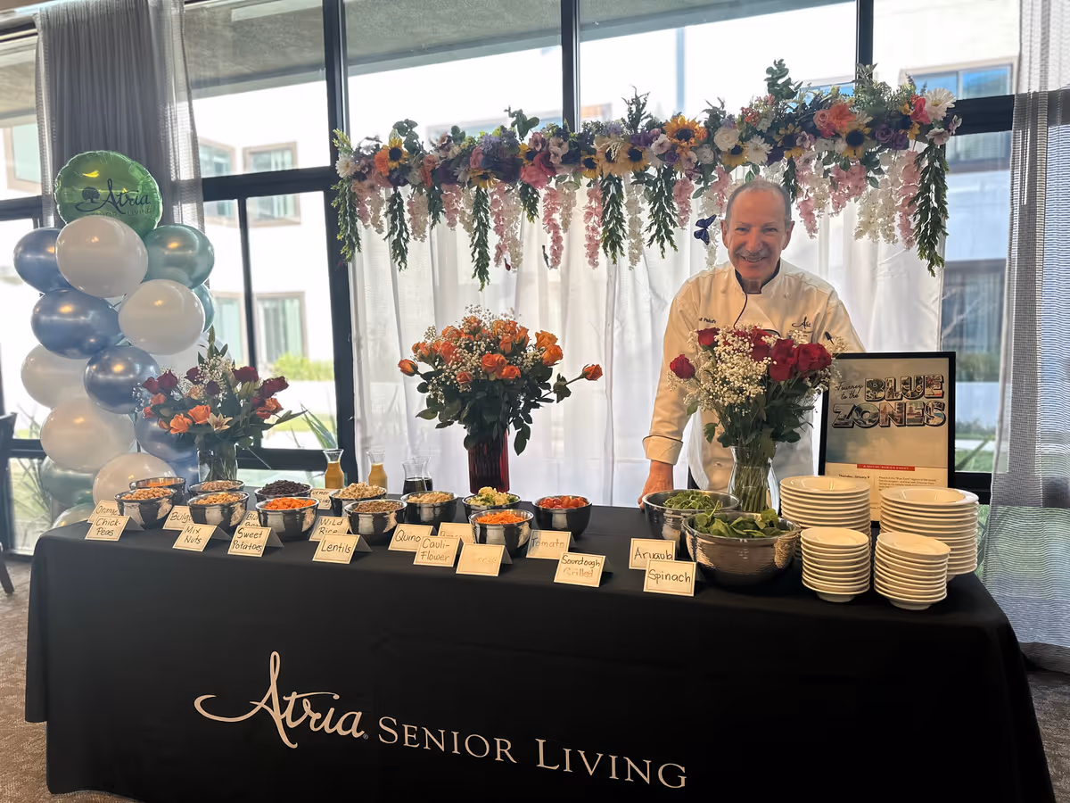 A chef stands behind a buffet table decorated with flowers and a balloon arrangement. The table has various bowls of food labeled with items like Orange Chick Peas, Bulgur Mix Nuts, Sweet Potatoes, Wild Rice, Lentils, Quinoa Cauliflower, Tomato, Sourdough Grilled, and Arugula Spinach. There are stacks of plates on the right side of the table and a framed sign about Blue Zones. The backdrop includes large windows and a floral garland hanging above.