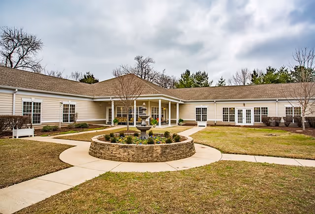 Exterior view of a single-story senior living facility building with beige siding and a brown roof, surrounding a circular stone planter with a tiered water fountain in the center. The area has paved walkways, some grass, and leafless trees under a cloudy sky.