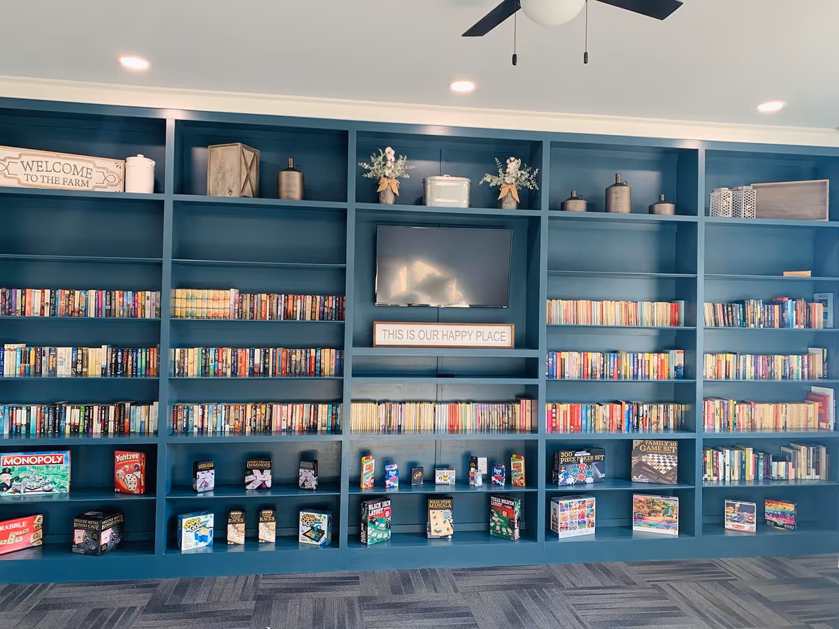 A large blue bookshelf filled with numerous books and board games. The bookshelf also has decorative items on the top shelves and a flat-screen TV mounted in the center with a sign below it that reads 'THIS IS OUR HAPPY PLACE'. The floor has a patterned carpet, and the ceiling has recessed lighting and a ceiling fan.