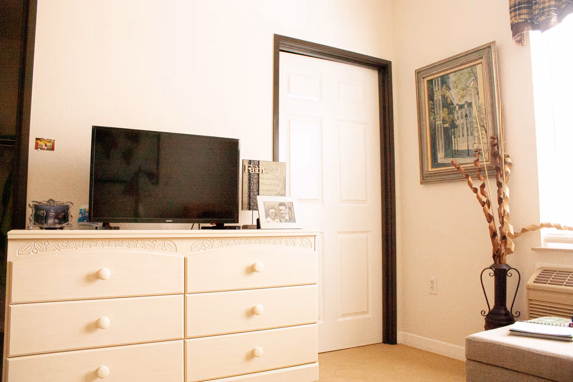 A bright room with a cream-colored dresser holding a flat-screen TV, framed photos, and decorative items. There is a white door with dark trim, a framed painting on the wall, a window with a plaid valance, and a vase with tall dried branches next to an air conditioning unit.