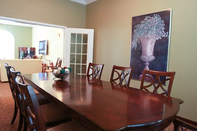 A dining room with a long polished wooden table surrounded by six wooden chairs with cushioned seats. On the table is a decorative bowl filled with assorted decorative balls. The room has beige walls with a large framed painting of a vase with flowers hanging on one wall. In the background, there is an adjoining living area with a sofa and a television mounted on the wall.
