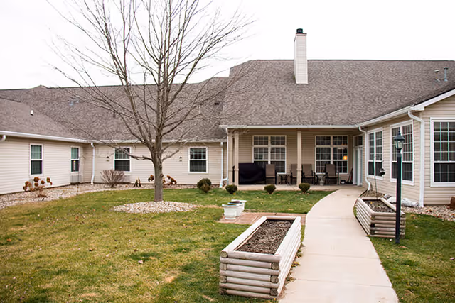A landscaped courtyard with a concrete walkway, raised planter boxes, a leafless tree, and a covered patio attached to a beige single-story building.