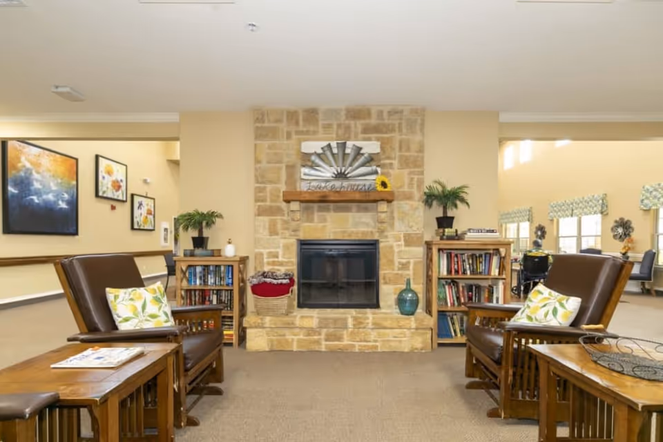 A cozy assisted-living common room with leather chairs, side tables, bookshelves and a stone fireplace.